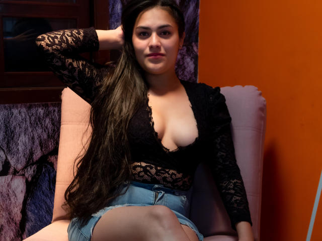 ChloeDumont with long brown hair, wearing a black lace top and denim shorts, sitting in a pink chair, hand behind head, showing her tits and a subtle smile.