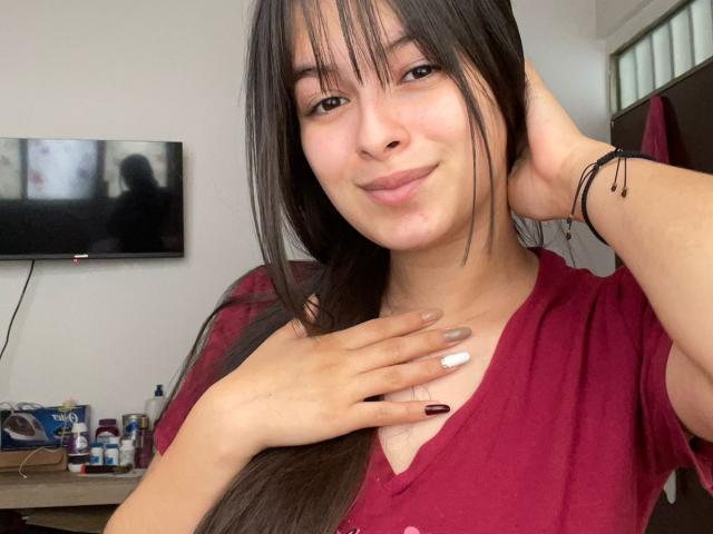 Kaary69 with long black hair, wearing a red shirt, hand on chest, smiling, has a white nail, dark nail on other hand, black bracelet, flat chest, light skin.