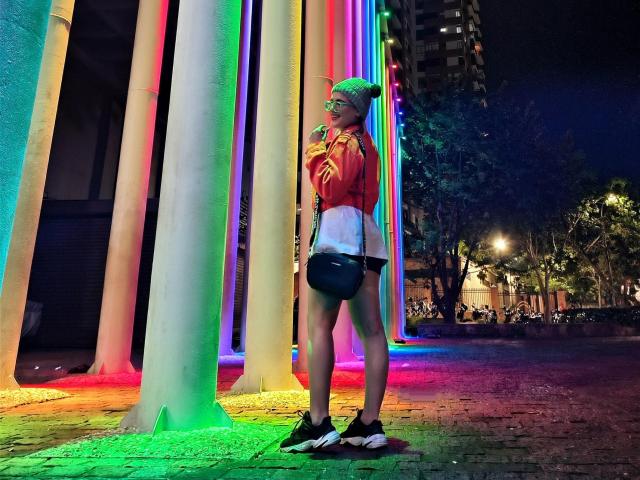 KimSummer se tient dans une zone éclairée au néon, portant une veste rouge, une chemise blanche, un short noir et des baskets, avec une casquette colorée, et regarde par-dessus son épaule, montrant une jolie fesse..