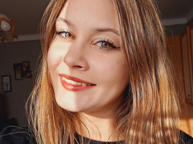 ValeriaDream has light brown hair, green eyes, and a cute smile with shiny red lipstick. Sunlight highlights her face, showing her long lashes and slightly wavy hair.