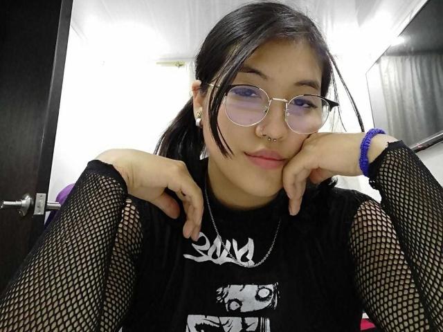 Elizaa, con coletas negras, gafas, aro en la nariz, camiseta de malla negra, manos en las mejillas, sonriendo, camiseta negra, pulsera azul, pendientes blancos, busto medio, puerta negra en el fondo..