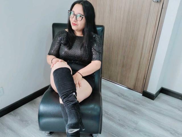 AnnabellaKinky mit schwarzen Haaren, Brille, einem durchsichtigen schwarzen Oberteil und schwarzen, hohen Stiefeln, sitzt auf einem schwarzen Ledersessel und zeigt ihr nacktes Gesäß und ihre helle Haut..