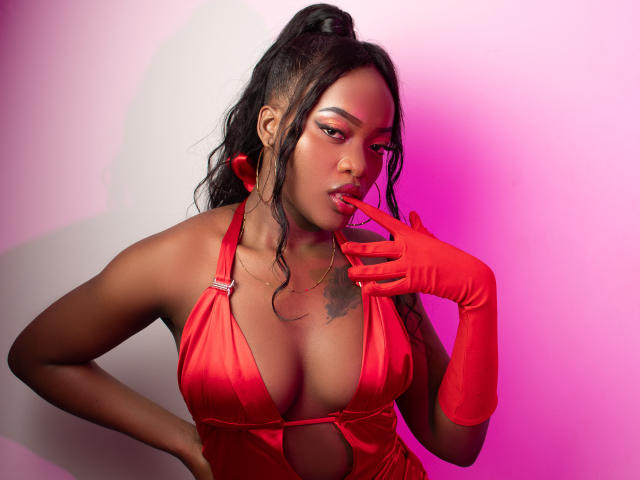 ZenyaEbony with dark skin, wearing a red satin halter dress, red gloves, and a high ponytail, sucking on her finger, showing her tits and tattoo on her chest.