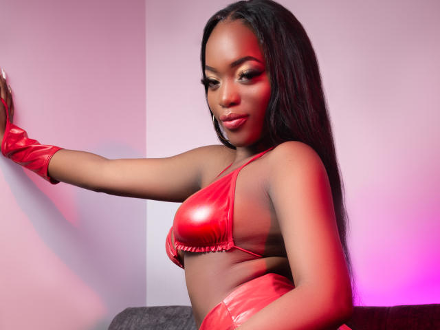 ZenyaEbony with dark skin and long black hair, wearing a shiny red latex bra and matching gloves, touching a wall with a pink and red light glow.