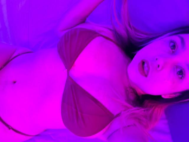AmayaJhons heeft roze en paarse borsten, draagt een bruine bikini-top, ligt plat met haar mond lichtjes open, heeft lang blond haar en een zichtbare navel..