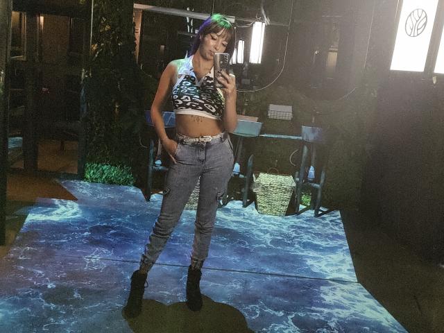 SamanthaMorgan with green hair, wearing a black-and-white patterned crop top, blue jeans, and black boots, standing in a studio, taking a selfie with hands in pockets.