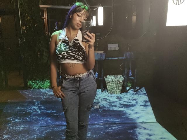 Samantha Morgan mit lila-grünen Haaren, die ein schwarz-weiß gemustertes Halstopp trägt, der ihre kleinen Brüste zeigt, und hoch taillierte Jeans, macht ein Selfie vor einem Spiegel in einem dunklen Raum..