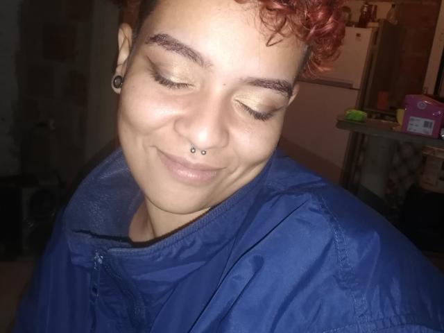 Shezka met kort, rood krullend haar, een neusring en een blauw jasje, lachend met gesloten ogen, waarbij haar Assy-neus en schattige gezicht zichtbaar zijn, en zwarte oorbelletjes draagt..