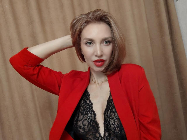 SimoniGrace con una giacca rossa, un reggiseno di pizzo nero, rossetto rosso, capelli castani corti, una mano tra i capelli, che guarda la fotocamera, una collana d'oro, uno sfondo con tende beige, che mostra il seno..