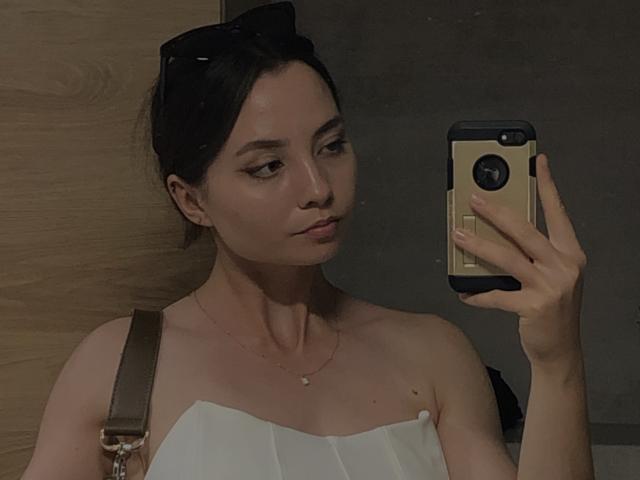 NicolePlay with dark hair, wearing black sunglasses on head, white strapless top, brown shoulder bag, holding phone, looking at mirror, small necklace, subtle makeup, dark room.
