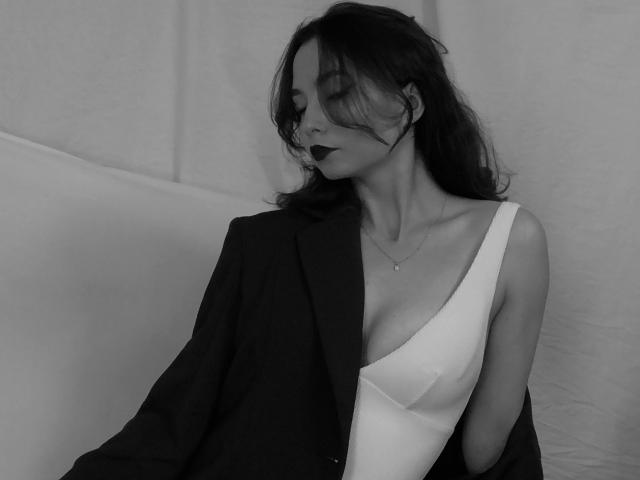 NicolePlay with dark lipstick, wearing a low-cut white dress and black blazer, has her eyes closed and hair partially covering her face, looking sexy and seductive.