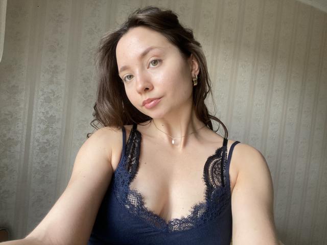 NicolePlay with medium tits in a navy lace top, light skin, brown wavy hair, subtle makeup, small nose ring, and necklace, looking at camera with a slight smile.