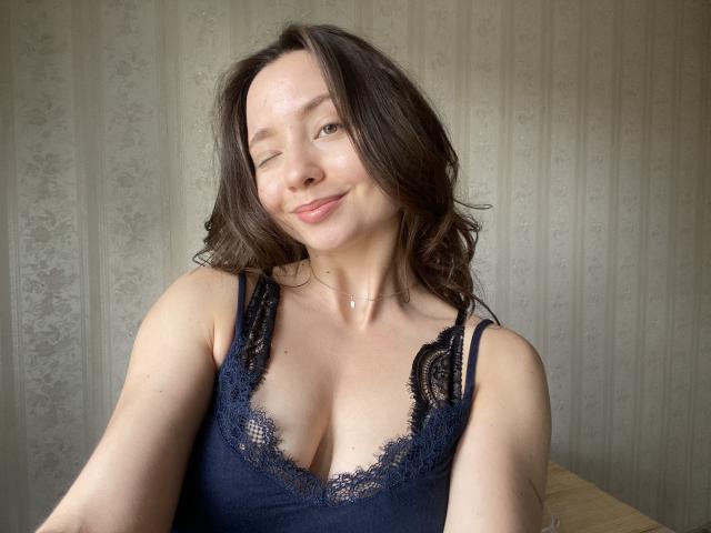 NicolePlay with wavy brown hair, one-eye wink, and subtle smile, wearing a dark blue lace camisole, showing her tits in the shot. Light skin, cute face.