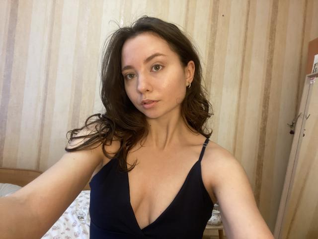 NicolePlay with wavy brown hair, wearing a black V-neck top, showing her tits, light skin, green eyes, and small earrings, taking a selfie in a bedroom.