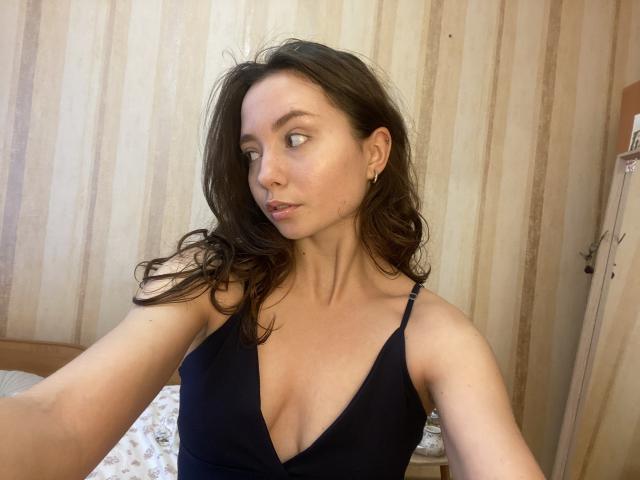 NicolePlay with wavy brown hair, wearing a black V-neck top, showing small tits and a slim build, looking to the side with a slight smile, in a bedroom.