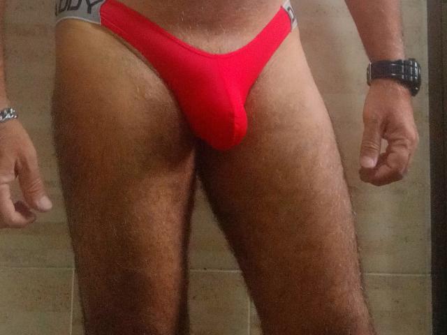 A photograph of CoupleMature's lower torso showing a red thong, highlighting their flaccid, bulging cock, with hairy thighs and a black wristwatch on their right wrist.