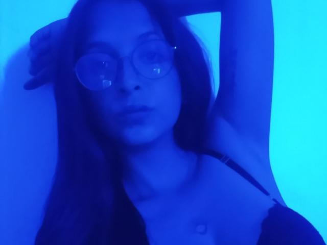 AmathystaBlack met een huid met blauwe tinten, een bril, lang donker haar en een zwarte top, die haar arm omhoog steekt en haar borsten laat zien, op een semi-erotische, sfeervolle foto..
