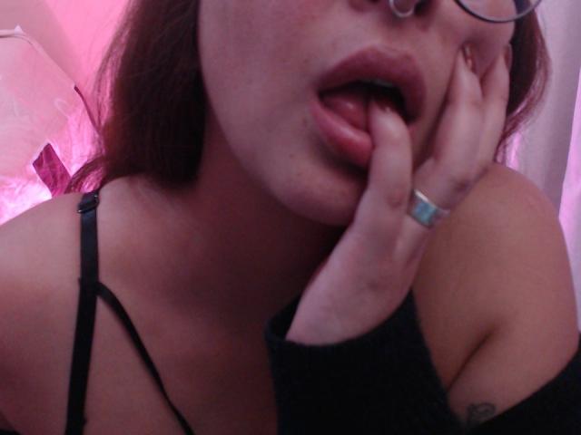 Il viso di AmathystaBlack, bocca leggermente aperta, lingua che tocca il labbro superiore, mano vicina alla bocca con un anello, occhiali, cinturino nero, illuminazione rosata, tatuaggio visibile sulla spalla destra..
