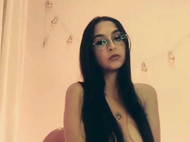 AmathystaBlack, con i suoi lunghi capelli neri, gli occhiali e la collana, ha i seni leggermente visibili e un'espressione pensierosa sul viso. Nella foto, non indossa una camicia..
