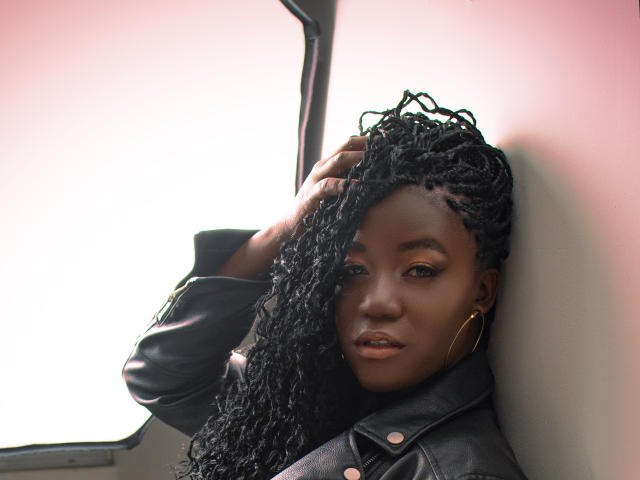 DianethKiss, peau foncée, longs cheveux noirs bouclés, portant une veste en cuir noir, boucles d'oreilles dorées, appuyée contre un mur, une main dans les cheveux, regardant la caméra, expression sérieuse..