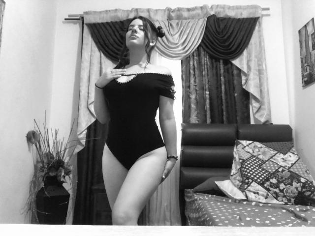 EmmaMack, avec de grandes seins, portant une combinaison noire à une épaule, debout, une main sur la poitrine, regardant vers le haut, sur une photo en noir et blanc d'une chambre..