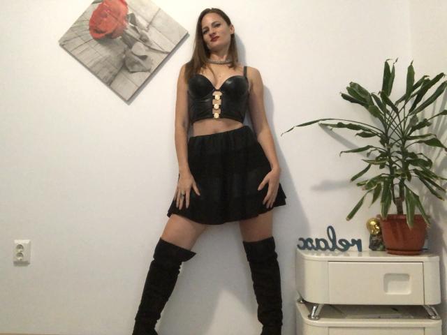 AbyX with long brown hair, black leather bustier showing her tits, black skirt, and thigh-high boots, standing with hands on her skirt, and a green plant to the right.