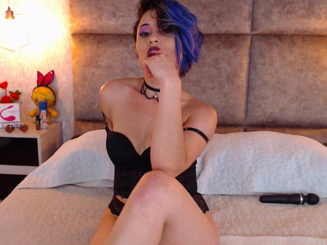CassieJones with blue hair, black lace top, and choker, sitting on bed, hand on cheek, looking at camera, legs crossed, black vibrator on bed, purple lipstick.