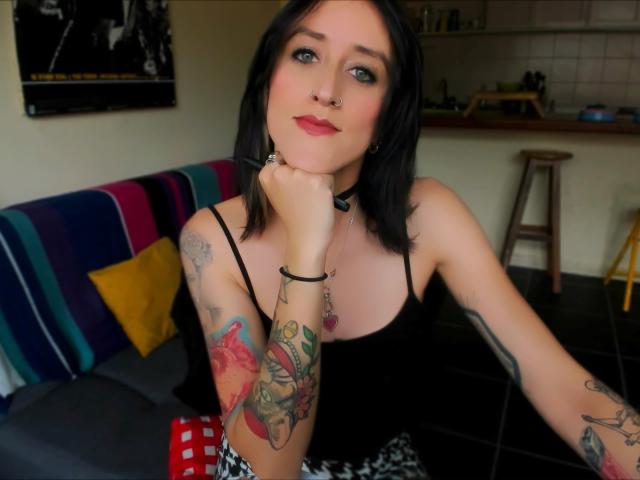 Envyv mit schwarzen Haaren, blauen Augen und Tätowierungen, trägt ein schwarzes Oberteil, einen Nasenring und eine Halskette, legt ihr Kinn in die Hand und zeigt ihre farbenfrohen Armtätowierungen..