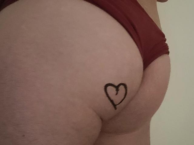 Oli69's butt in red panties with a small black heart tattoo on the right cheek. The tattoo is simple and centered. Skin is light. Photo is slightly blurry.
