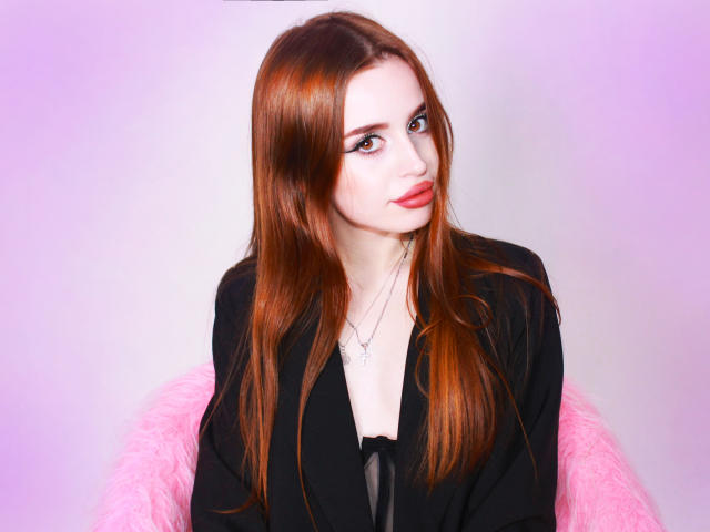 CherylFoxy has long red hair, fair skin, and red lipstick. She's wearing a black top with a silver necklace, sitting on a pink fuzzy chair.