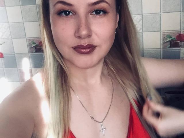 HanterHellen, con i capelli biondi, una maglietta rossa e una collana con una croce, si tiene i capelli, guarda la fotocamera, indossa un rossetto rosso scuro, in un bagno molto luminoso..