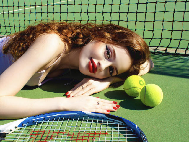 CherylFoxy with red lipstick, lying on tennis court, hands on racket, red nails, yellow balls, curly brown hair, green background, looking at camera, wearing white top.