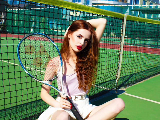 CherylFoxy with long red hair, sitting on a tennis court, holding a racket, wearing a white dress, and red lipstick, with a net in the background. Boobs visible.