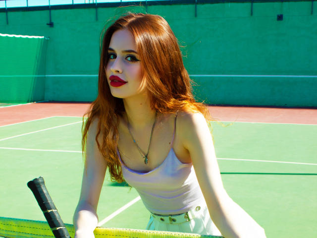 CherylFoxy with red hair, white tank top, and yellow skirt, holding a tennis racket, standing on a green tennis court, wearing red lipstick, and a necklace.