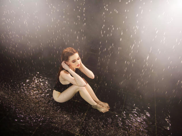 CherylFoxy, wet, sitting on dark floor, wearing black one-piece, hands on head, rain pouring down, water on skin, bare feet, red hair, sad expression, legs crossed, water droplets.