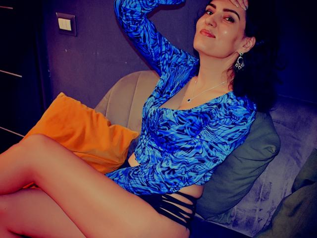 AddictedToUTS with blue patterned top, black strappy bottoms, and earrings, sitting on a gray couch, legs crossed, hand on head, looking up with a slight smile.