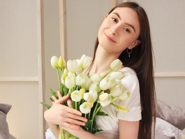 SaraQuincy, avec de longs cheveux bruns, une chemise blanche, tenant un bouquet de tulipes blanches, souriante, de petites boucles d'oreilles, peau claire, lèvres rosées délicates, regard doux, légère inclinaison, pose détendue..