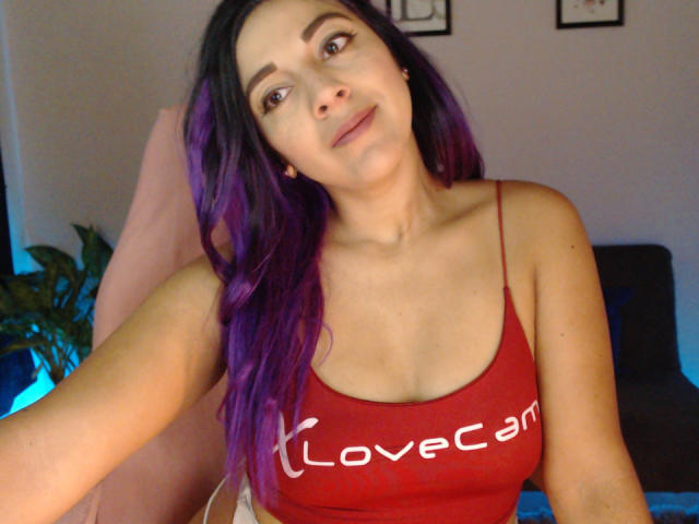 PrettyEllie with purple-dyed hair, red LoveCam tank top, showing her tits, slight smile, brown eyes, light skin, in a relaxed indoor setting, arm extended.