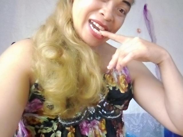 Rahel69 with blonde curls, smiling, finger in mouth, wearing a floral dress, black background, light skin, showing teeth, upper body, selfie, looking playful.