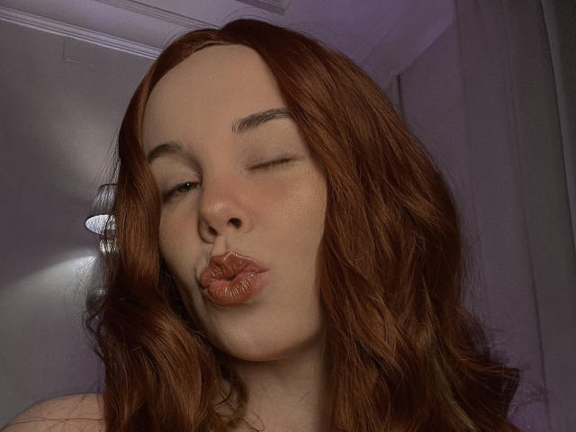 JaneRoller, avec ses lèvres boudeuses, ses yeux fermés et ses cheveux roux ondulés, fait une moue de baiser, montrant son nez et ses joues légèrement rougies. Ses seins ne sont pas visibles..