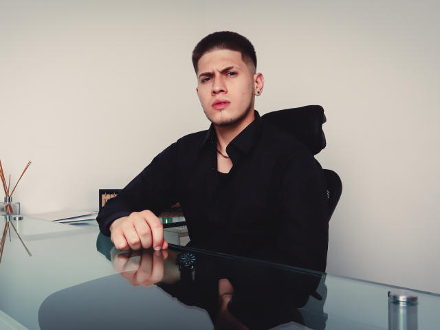 SebastiianMaxime, com cabelo castanho curto, moletom preto e expressão irritada, está apoiado em uma mesa de vidro, que reflete seu rosto, com um difusor de varetas ao fundo..