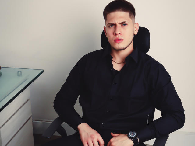 SebastiianMaxime with short brown hair, black shirt, and black watch, sitting in a chair, looking serious, hands on lap, glass table on left.