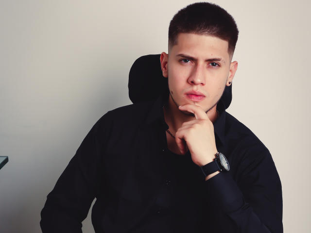 SebastiianMaxime mit kurzen, braunen Haaren, schwarzem Hemd und Armbanduhr, mit dem Kinn auf den Fingern ruhend, wirkt ernst, trägt kleine Ohrringe, vor einem schlichten weißen Hintergrund..