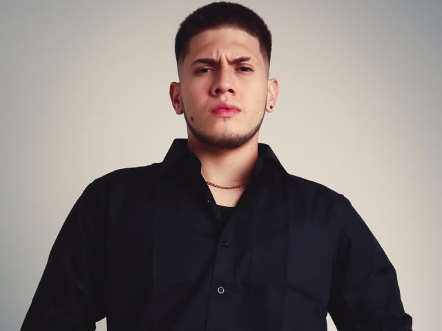 SebastiianMaxime ha i capelli castano corti, un piccolo pizzetto e un'espressione sofferente, indossa una camicia nera con una catena dorata e piccoli piercing alle orecchie..
