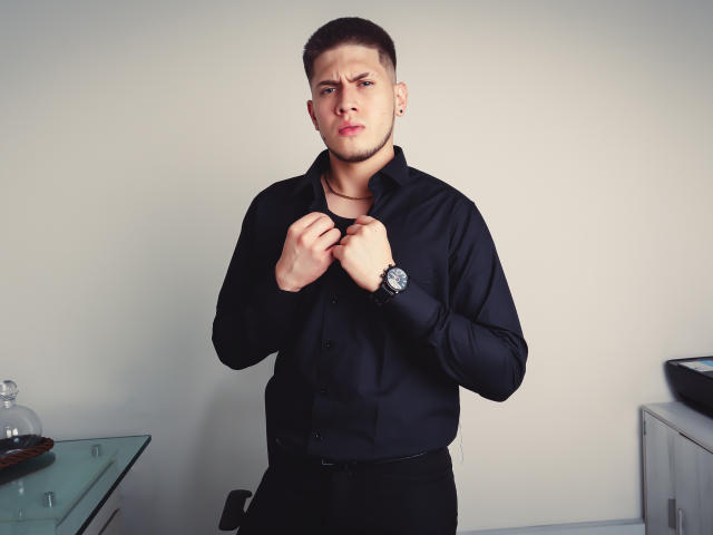 SebastiianMaxime, muscular, short brown hair, black shirt, buttoning up, serious expression, black pants, wristwatch, glass table with bottle, white wall, right side cabinet, metal bowl on top.