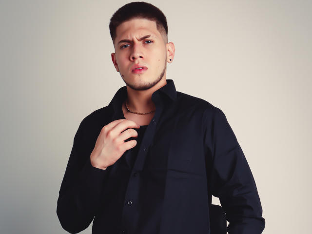 SebastiianMaxime, con cabello castaño corto, camisa negra y aretes, con expresión molesta, sosteniendo el cuello de la camisa con la mano derecha, piel clara, ligera barba incipiente, un pequeño aro en la nariz y un collar negro..