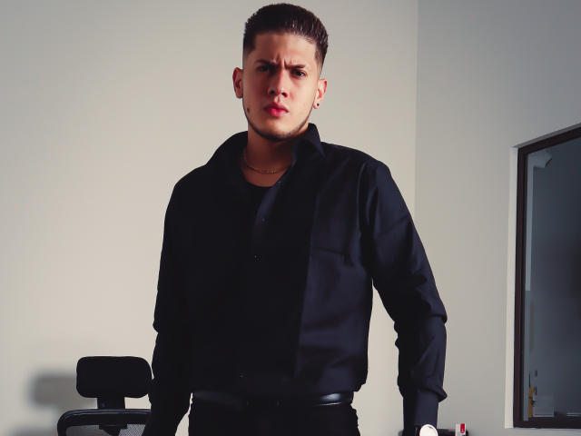 SebastiianMaxime steht mit ernstem Gesichtsausdruck da, trägt ein schwarzes Hemd und eine schwarze Hose, hat die Hände in schwarzen Handschuhen, kurze braune Haare und eine kleine Halskette, in einem schwach beleuchteten Raum..