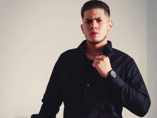 SebastiianMaxime, com cabelo castanho curto, camisa preta e relógio prateado, segurando a gola com uma expressão séria e ligeiramente irritada. Sua mão direita está perto do pescoço..