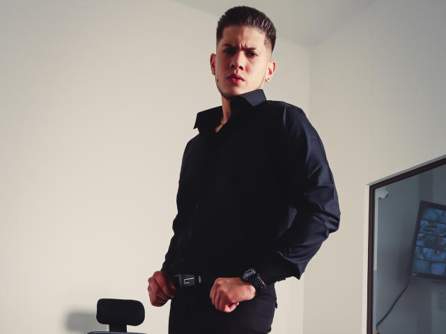 SebastiianMaxime está em pé, com uma expressão séria, vestindo uma camisa e calças pretas, com as mãos na cintura, em um quarto minimalista. Cabelo castanho curto, paredes brancas, cadeira preta..