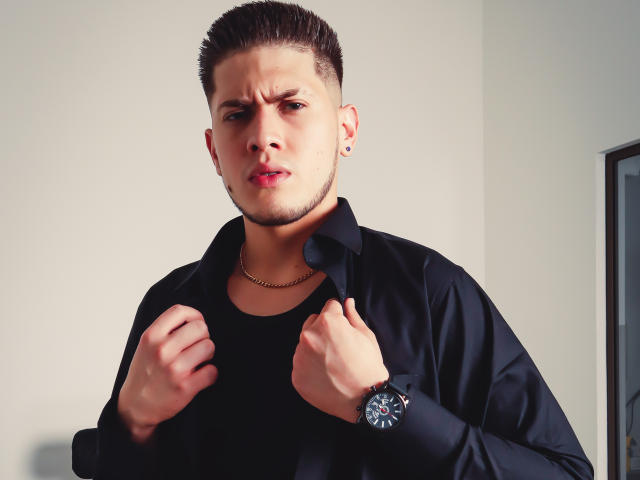 SebastiianMaxime, capelli castano corti, camicia nera, canottiera nera, catena d'oro, orologio da polso nero, con un'espressione corrucciata, che tiene i bordi della camicia, con piercing alle orecchie, pelle chiara, labbra rosse leggermente divisa, espressione seria..